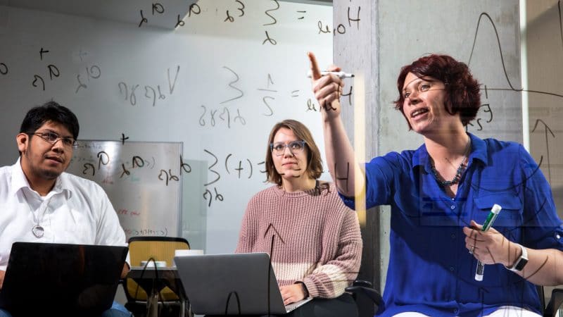 Faculty member pointing to an equation of a clear whiteboard while instructing two students with laptops