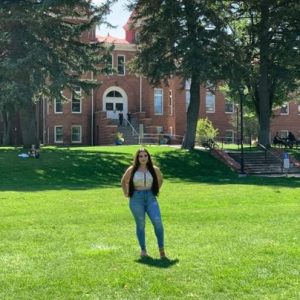 Photo of Elena Garcia standing in green grass in front of Old Main wearing jeans and a yellow shirt