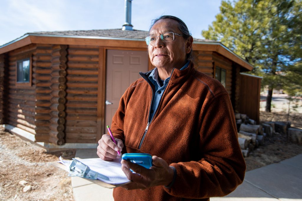 Martin stands in front of a log cabin and makes a note on a clipboard.