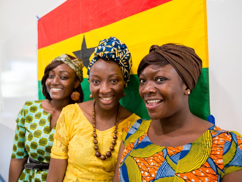Three students smiling in front of the flag of Ghana.
