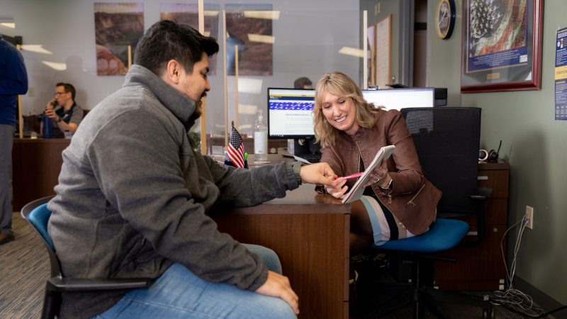 A student sits across from someone at a desk, and both are pointing at something on a notepad."